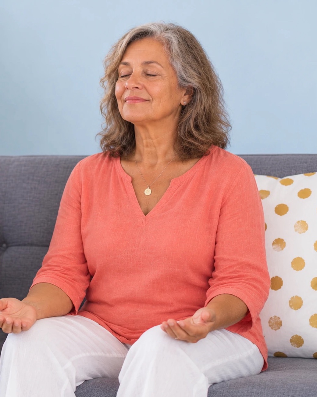 A woman sitting on a sofa, eyes closed and palms open, softly smiling
