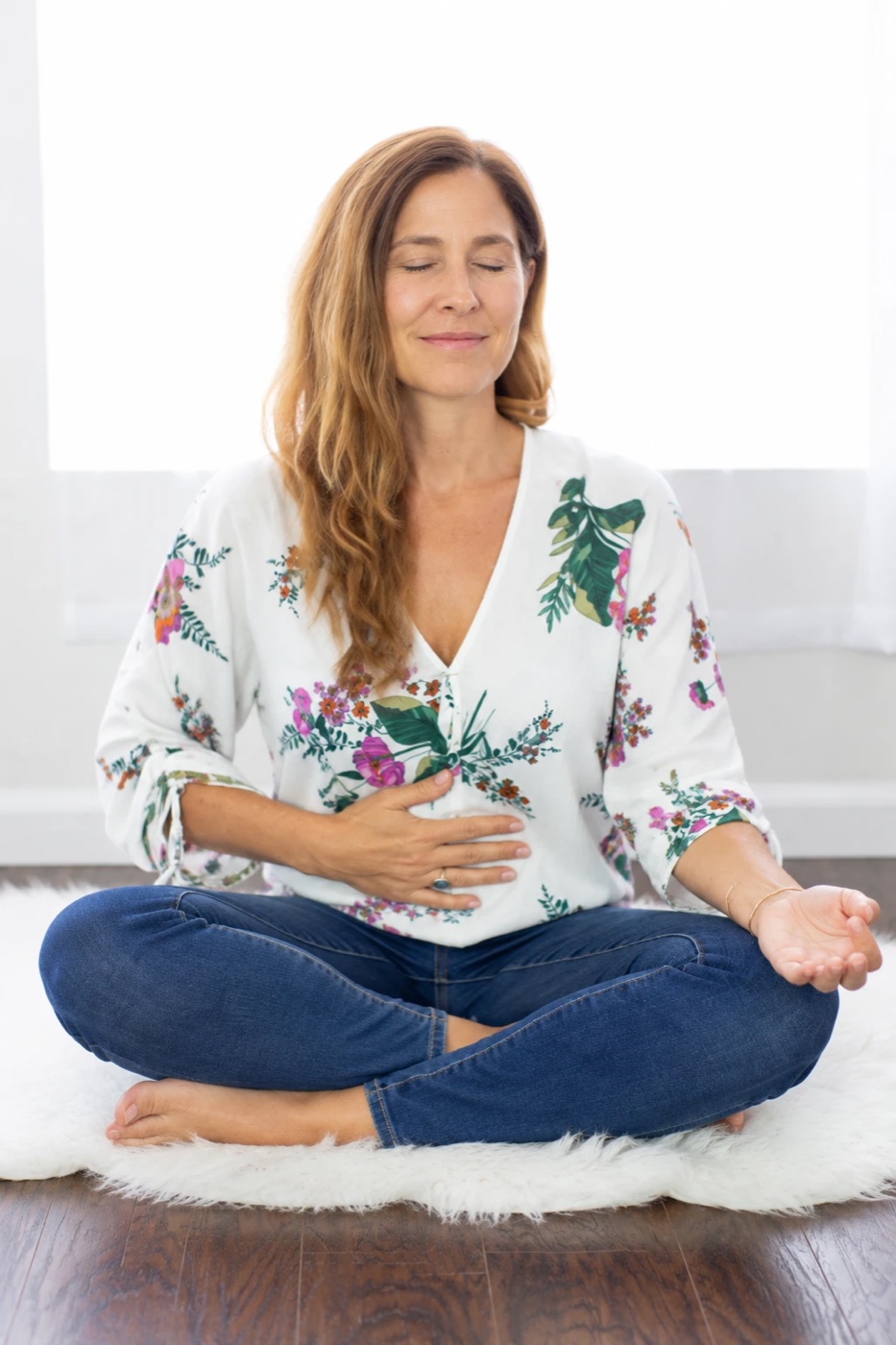 Sienna sitting cross-legged on a soft rug, eyes closed, one hand on her belly, breathing deeply