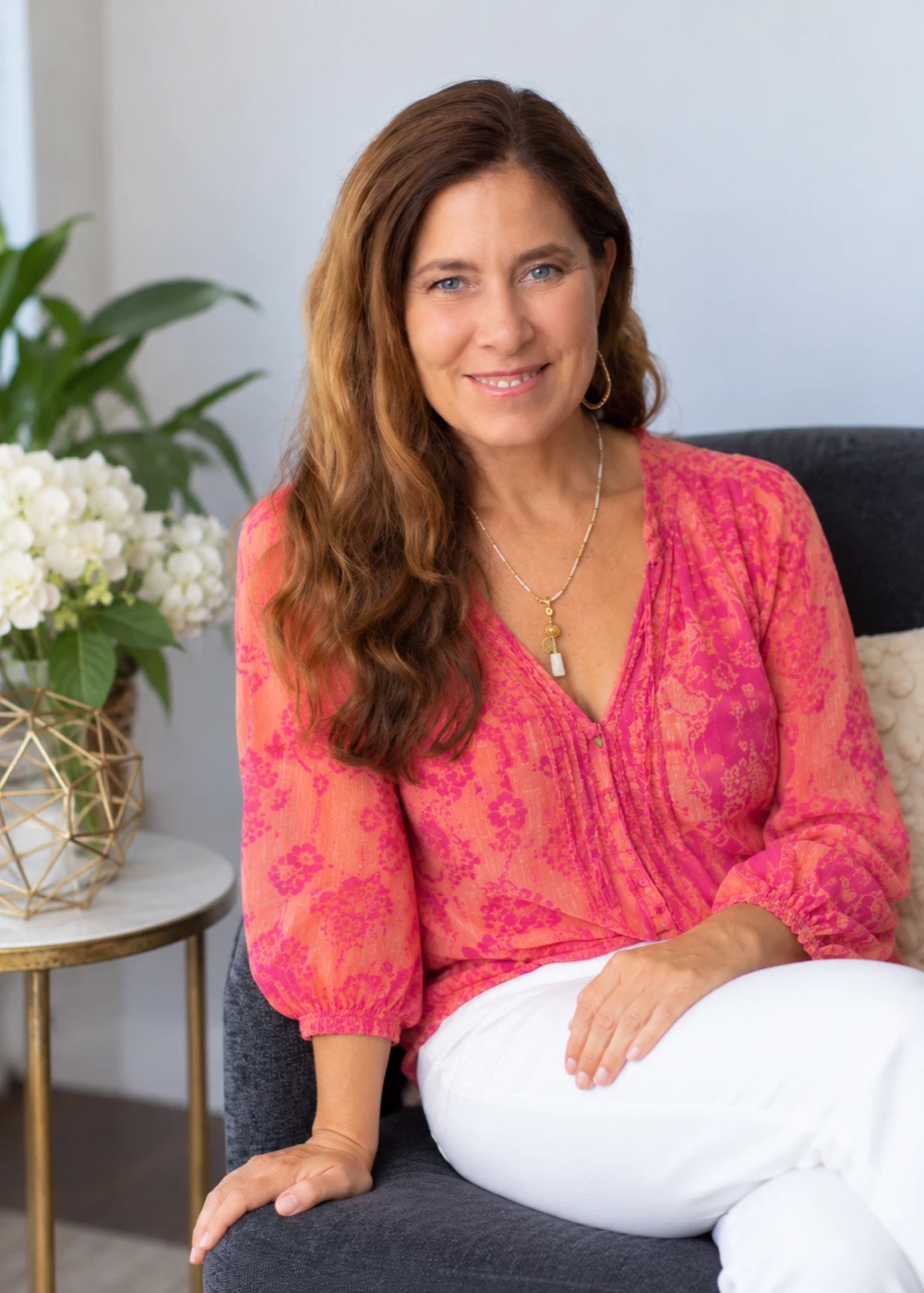 Sienna smiling, seated in a chair in a pink floral blouse beside white hydrangeas
