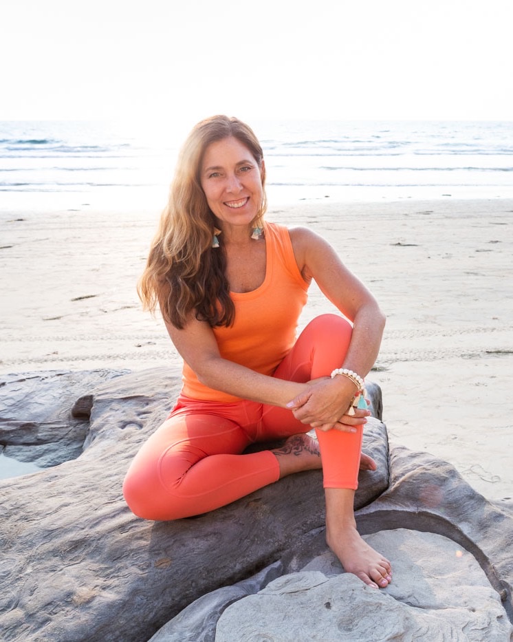 Sienna smiling, seated on a rock on the beach in orange yoga clothes
